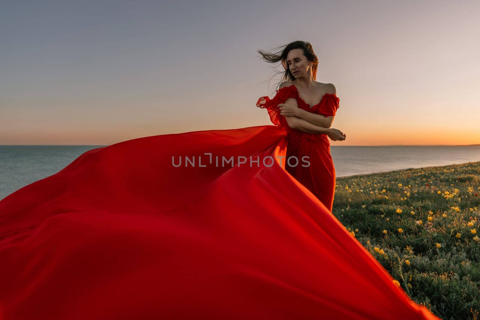 woman red dress standing grassy hillside. The sun is setting in the background, casting a warm glow over the scene. The woman is enjoying the beautiful view and the peaceful atmosphere