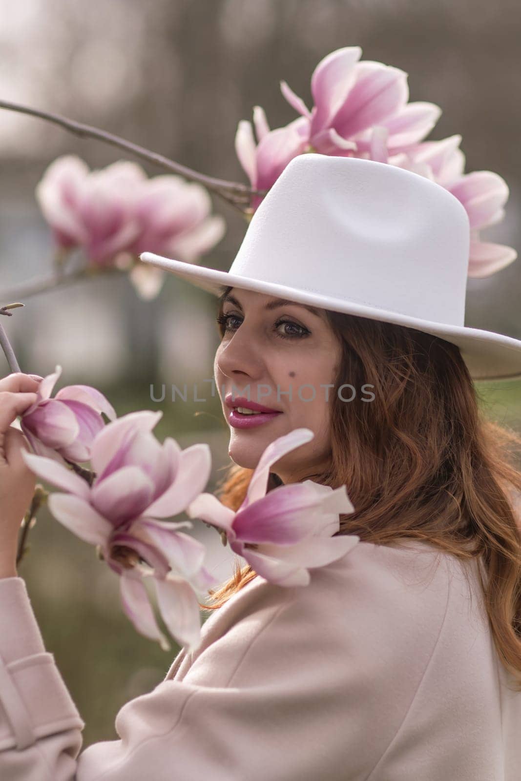 Woman magnolia flowers, surrounded by blossoming trees., hair down, white hat, wearing a light coat. Captured during spring, showcasing natural beauty and seasonal change. by Matiunina