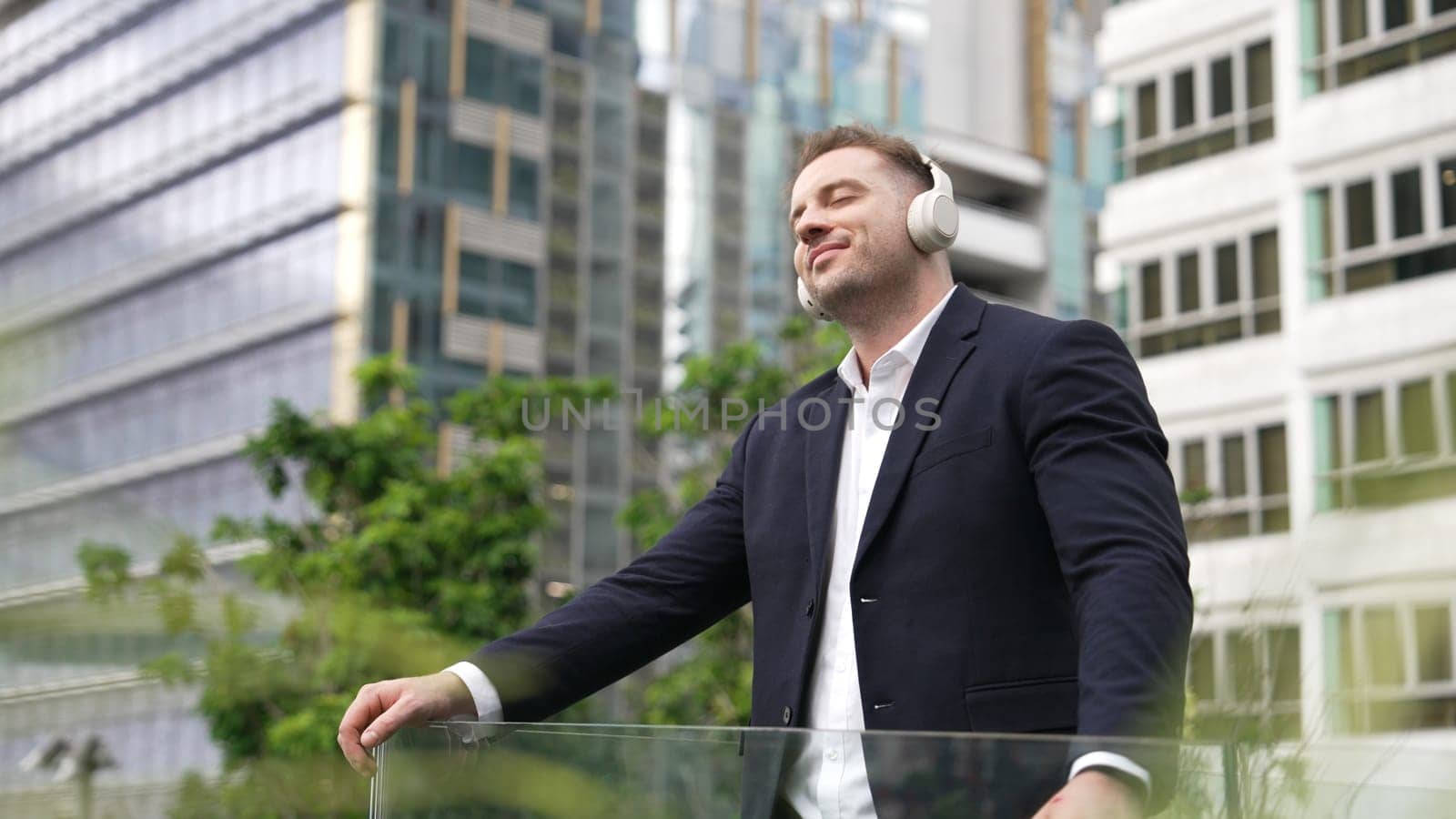Professional businessman wearing headphone while listening music at rooftop. Caucasian project manager moving to relaxing music while looking at skyscraper and stand at green city. Lifestyle. Urbane.