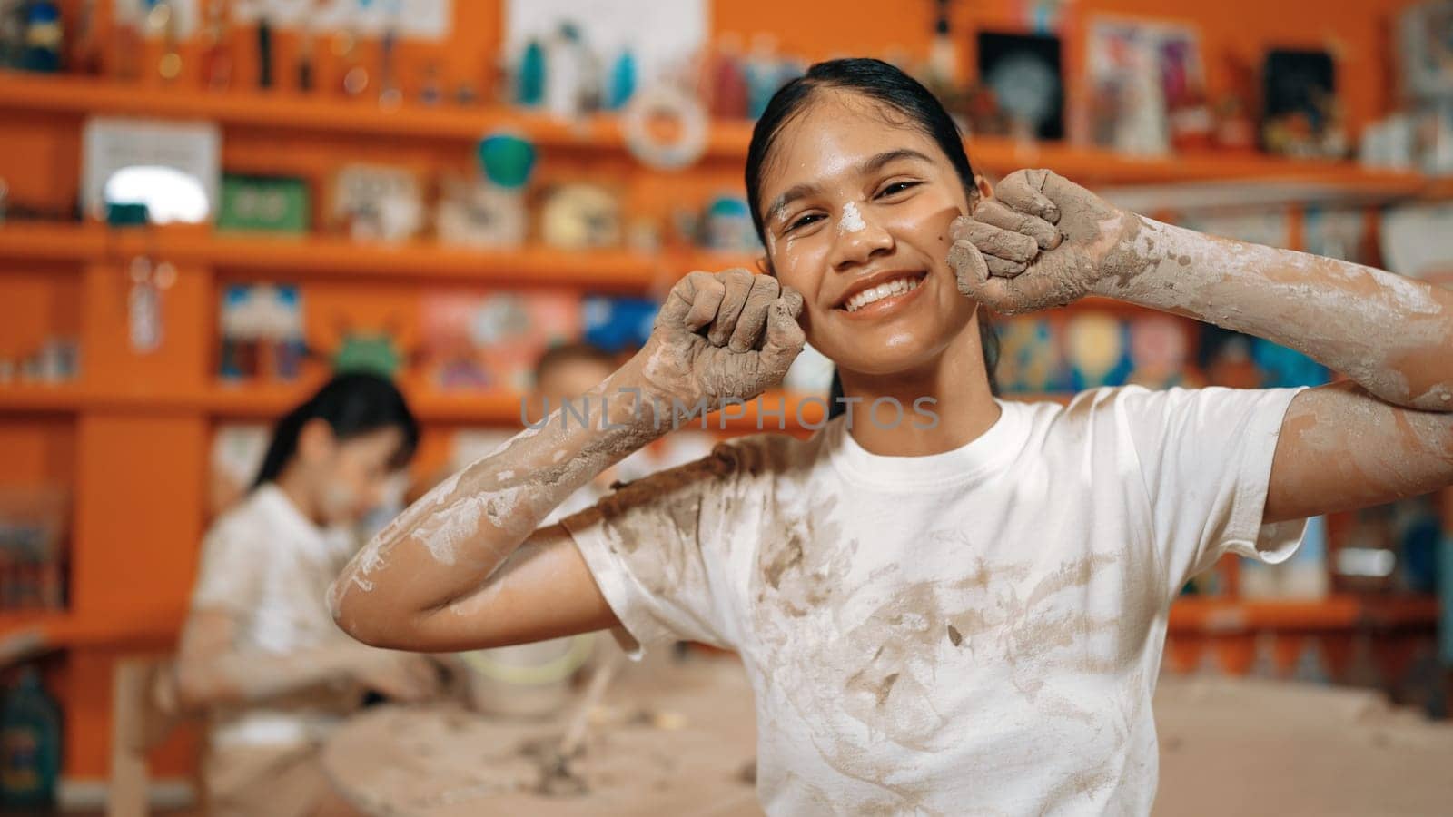 Happy caucasian girl pose at camera while diverse children modeling clay behind. Cute student wearing dirty shirt while looking at camera at workshop in art lesson. Blurring background. Edification.