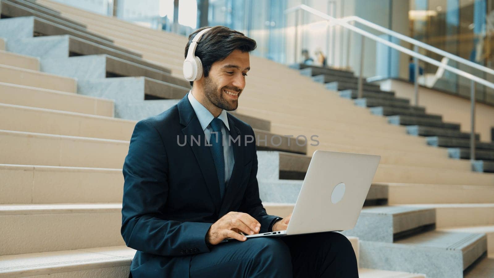 Professional business man sitting at stairs while working on laptop. Skilled project manager listening music from headphone and checking email and discussion about marketing plan. Outdoor. Exultant.