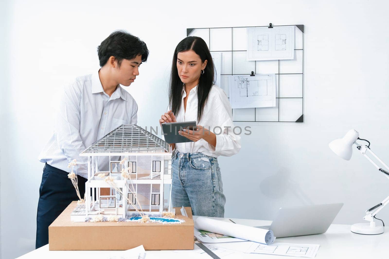 Beautiful young caucasian project manager tells asian engineer about building detail by using tablet while engineer measuring house model on meeting table with blueprint scatter around. Immaculate