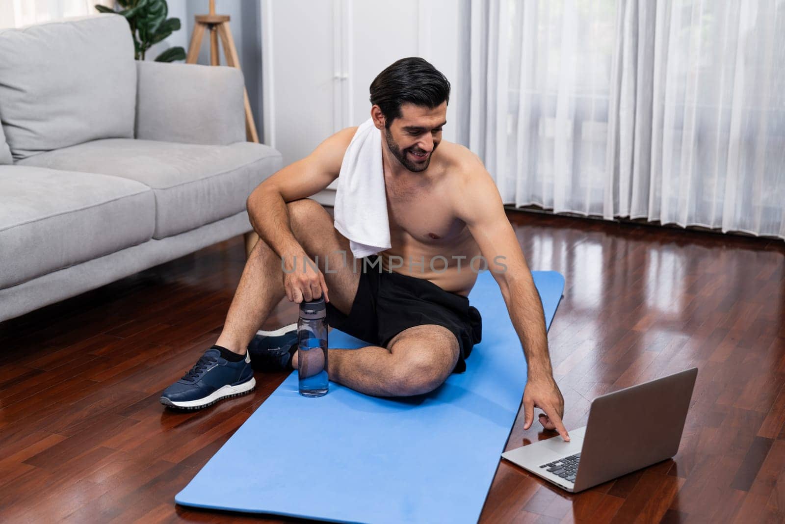 Athletic and sporty man resting on fitness mat during online body workout exercise session for fit physique and healthy sport lifestyle at home. Gaiety home exercise workout training concept.