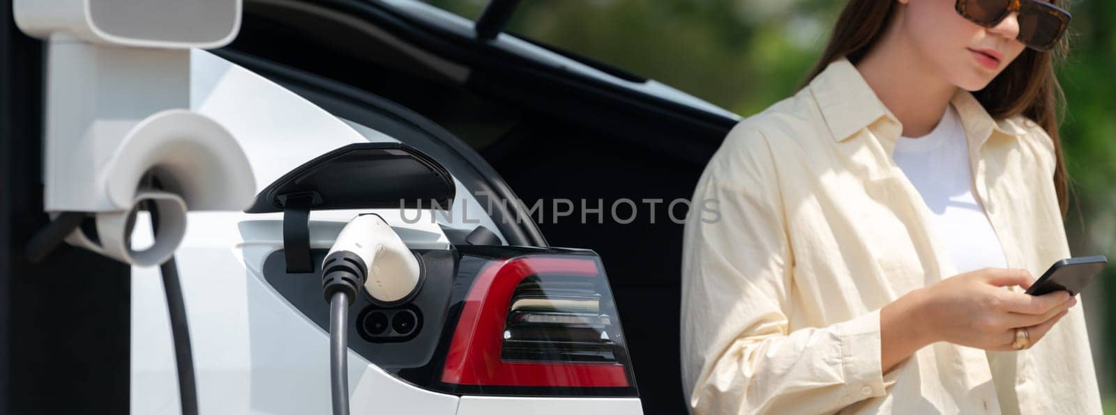 Young woman use smartphone to pay for electricity at public EV car charging station green city park. Modern environmental and sustainable urban lifestyle with EV vehicle. Panorama Expedient