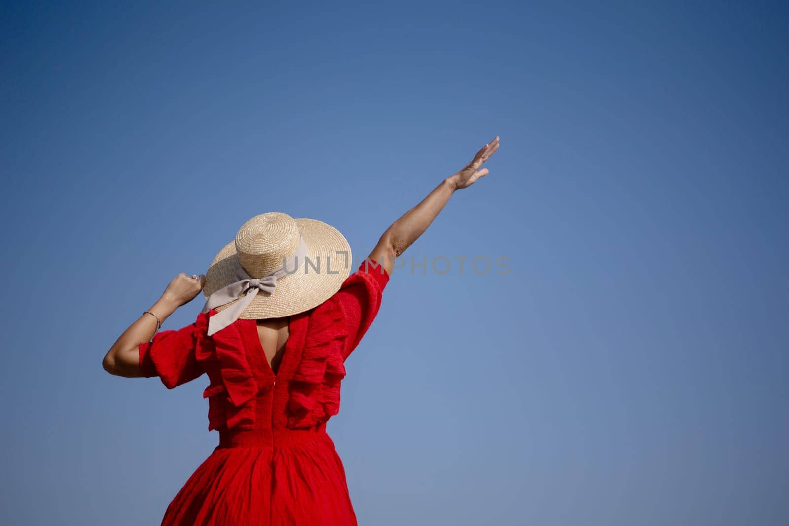 A woman in a red dress is standing in the sun with a straw hat on. She is waving her hand in the air