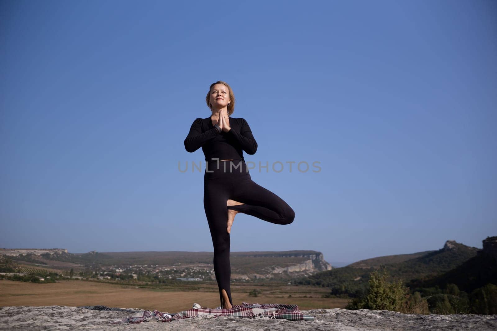 A woman is doing yoga on a rock in a beautiful landscape. She is in a pose that looks like she is balancing on one leg. The sky is clear and blue