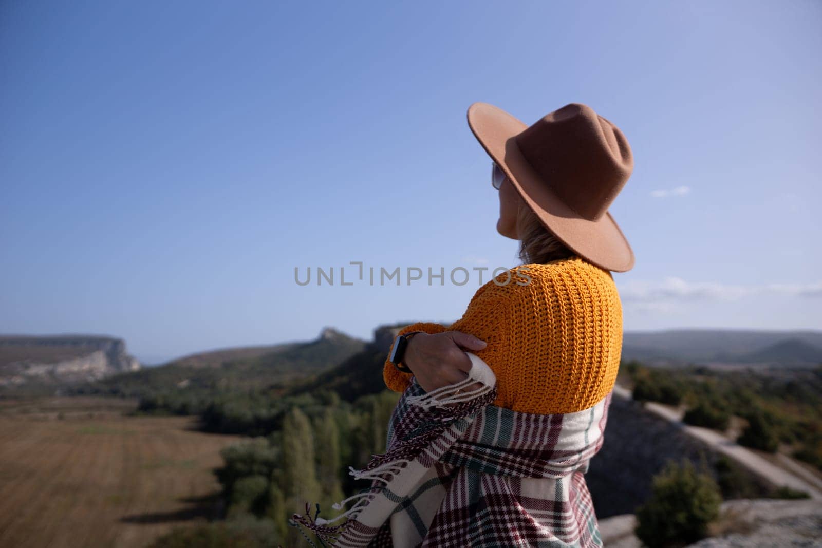 A woman wearing a brown hat and a yellow sweater is standing on a hillside, looking out at the landscape