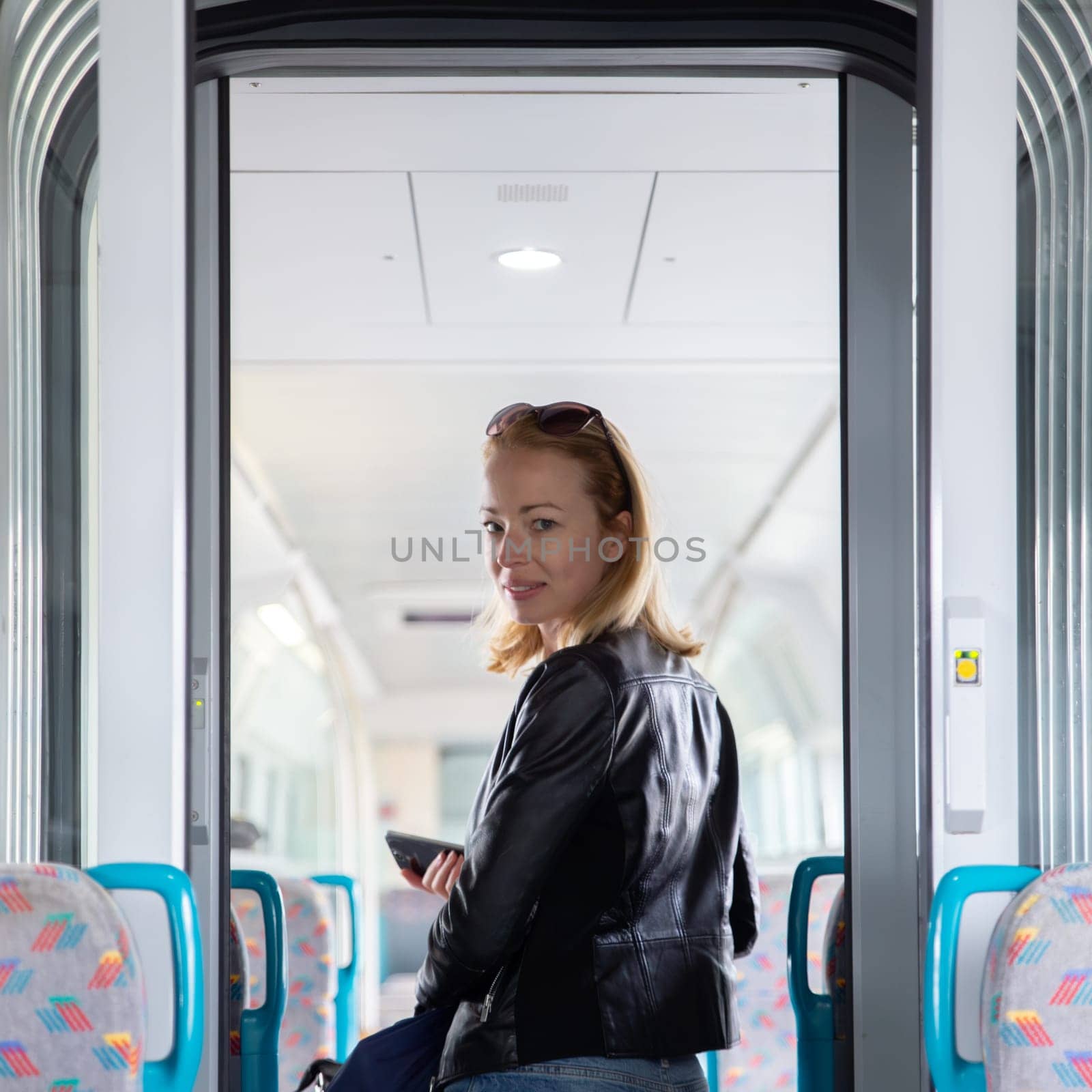 Young blond woman in jeans, shirt and leather jacket holding her smart phone and purse while riding modern speed train arriving to final train station stop. Travel and transportation. by kasto