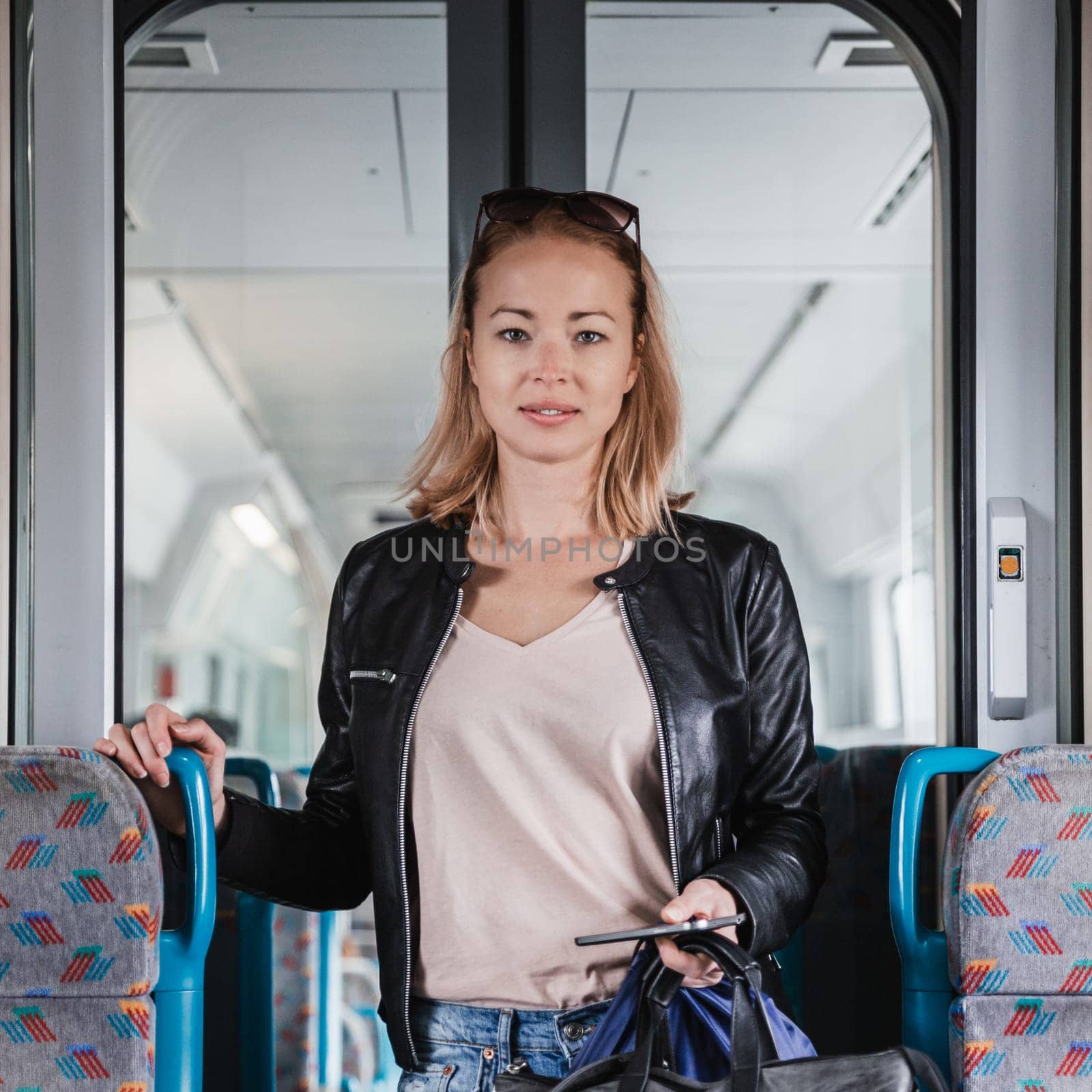 Young blond woman in jeans, shirt and leather jacket holding her smart phone and purse while riding modern speed train arriving to final train station stop. Travel and transportation. by kasto