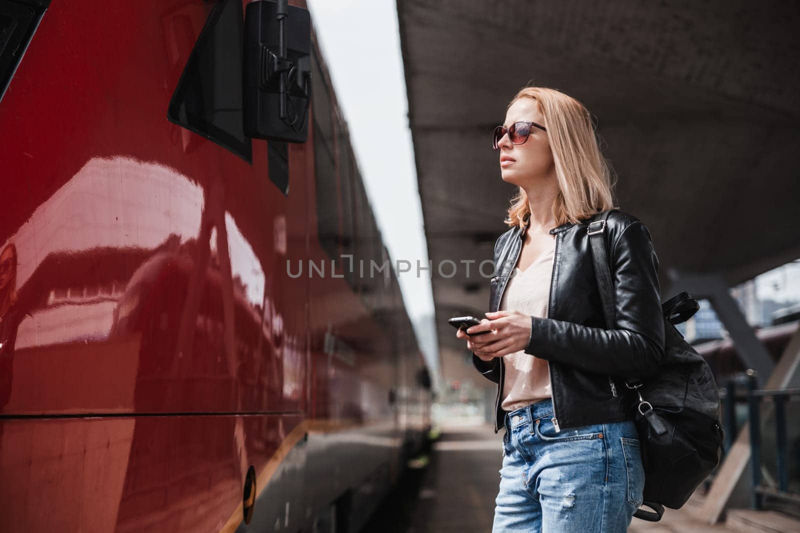 Young blond woman in jeans, shirt and leather jacket wearing bag and sunglass, embarking red modern speed train on train station platform. Travel and transportation