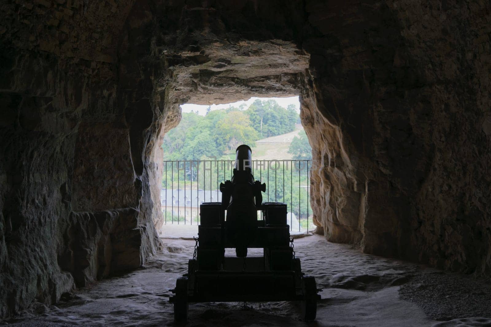 A silhouette of an old cannon inside a cave, facing an opening with a view of trees and a building in the distance.