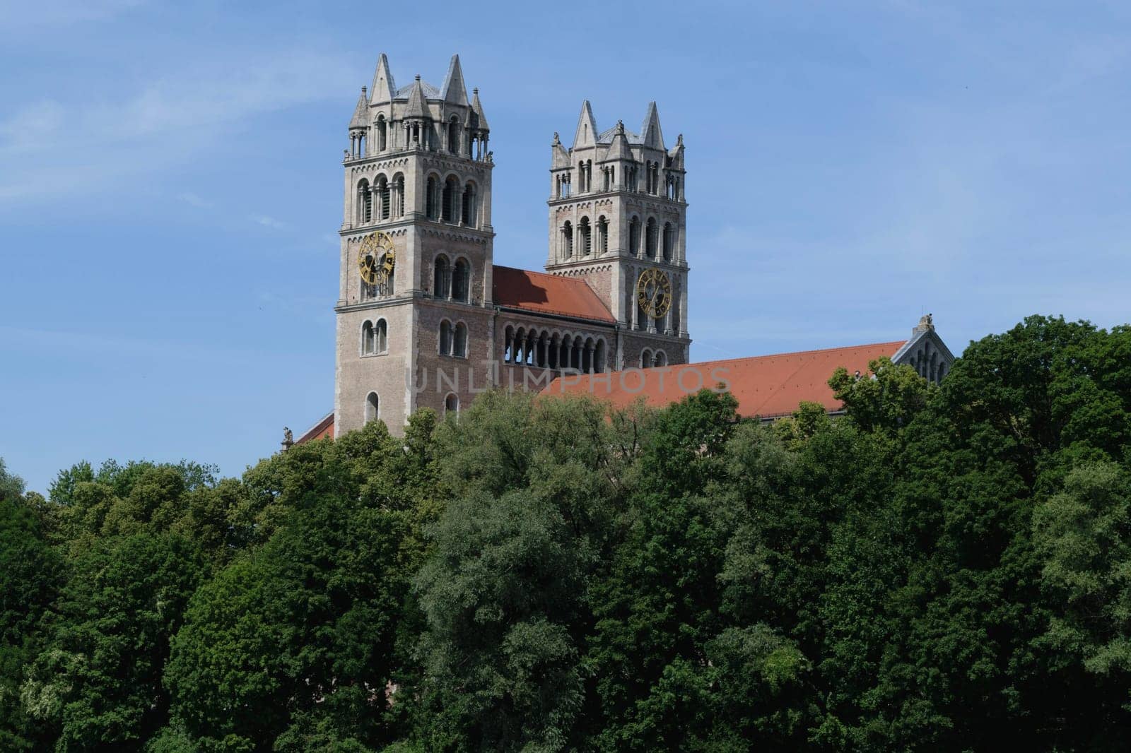 A historic church with twin towers and a red roof, partially obscured by lush green trees, under a clear blue sky.