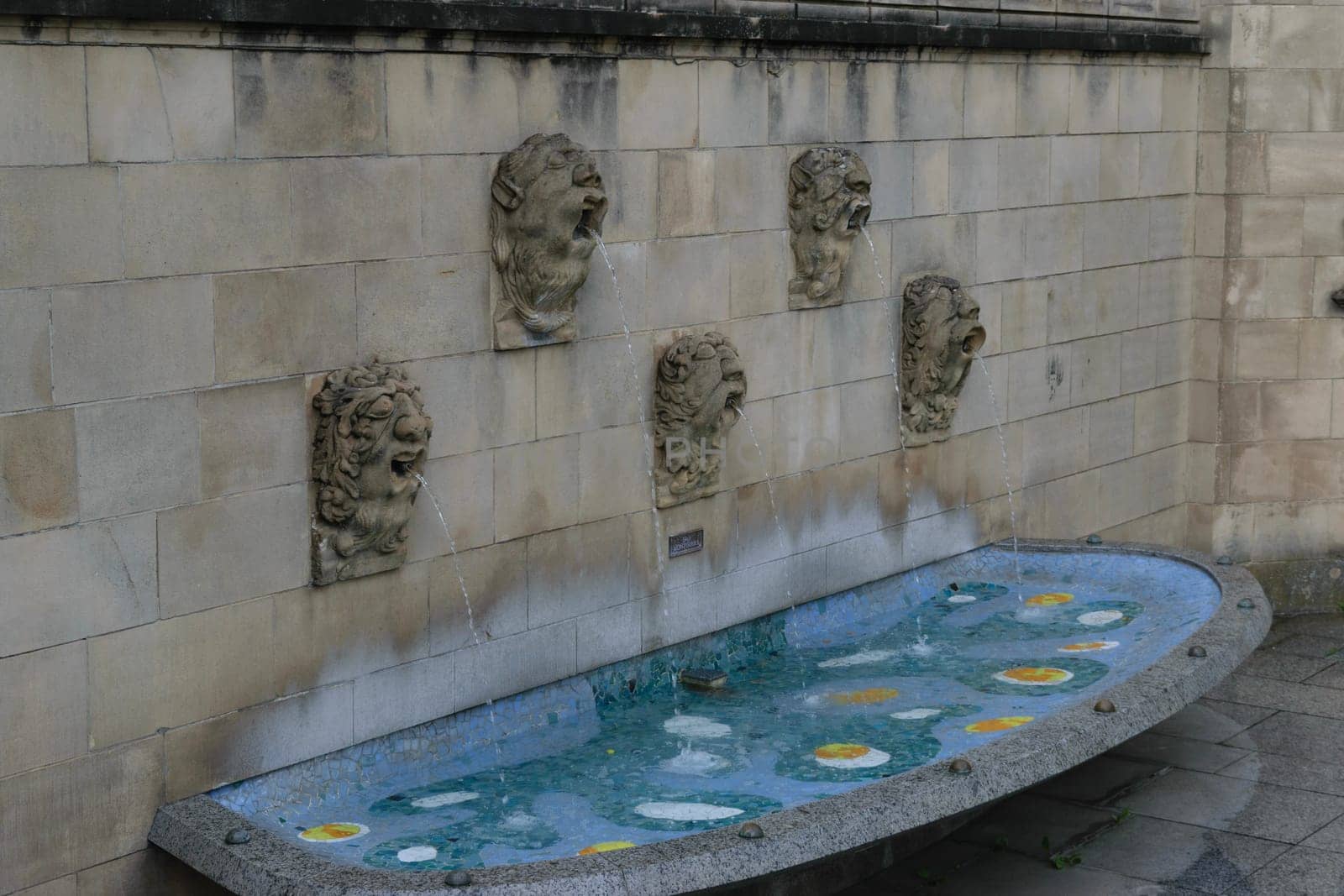 A stone wall with four lion head sculptures spouting water into a shallow, oval-shaped basin with a mosaic of yellow and white flowers.