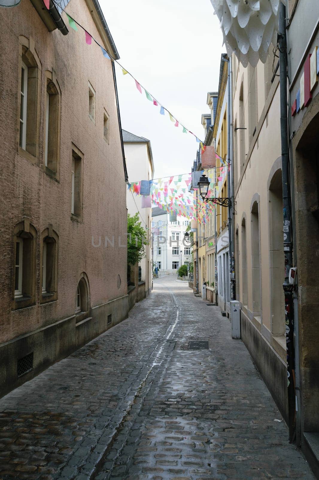 A narrow cobblestone street, adorned with colorful hanging flags. The street is lined with old buildings with arched doorways and windows.