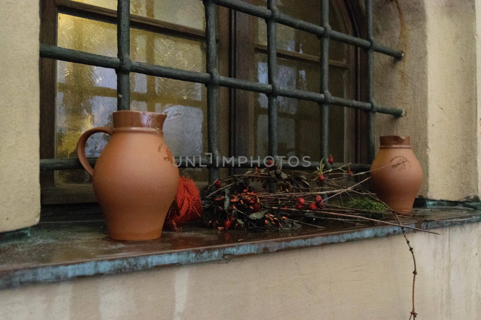 Two ceramic jugs and a bundle of dried branches with berries placed on a windowsill with metal bars. The window has an arched top and is set in a rustic, weathered wall.