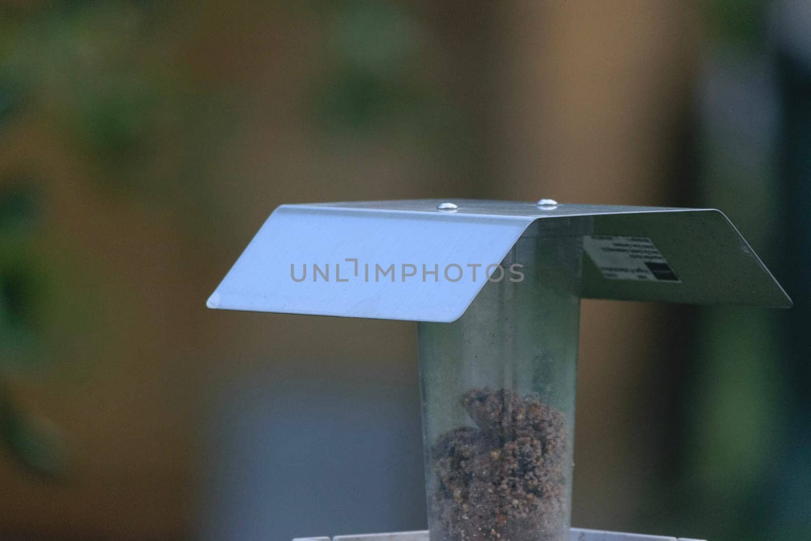 A close-up of a bird feeder with a metal roof and seed inside, set against a blurred natural background.