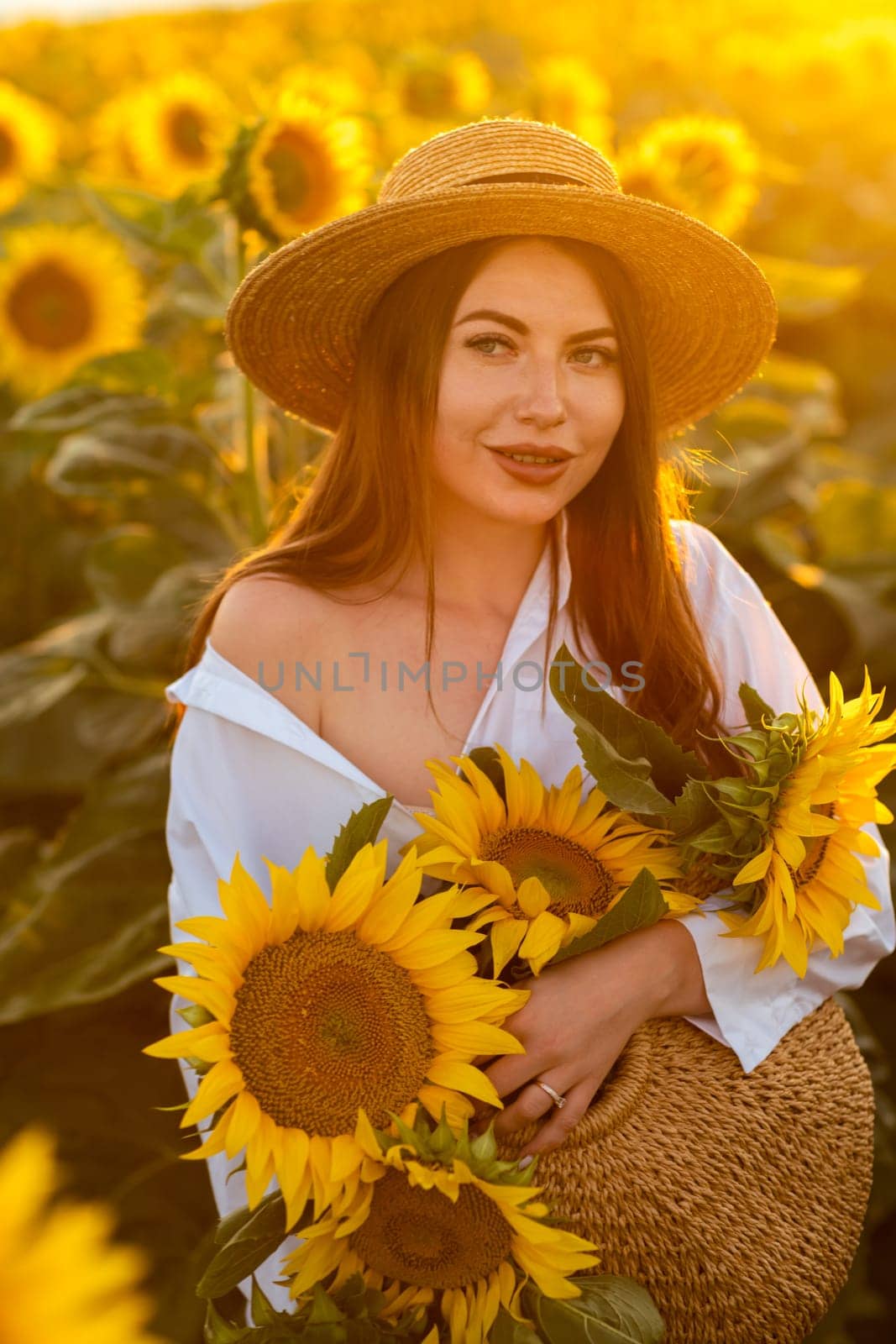 A girl in a hat on a beautiful field of sunflowers against the sky in the evening light of a summer sunset. Sunbeams through the flower field. Natural background. by Matiunina