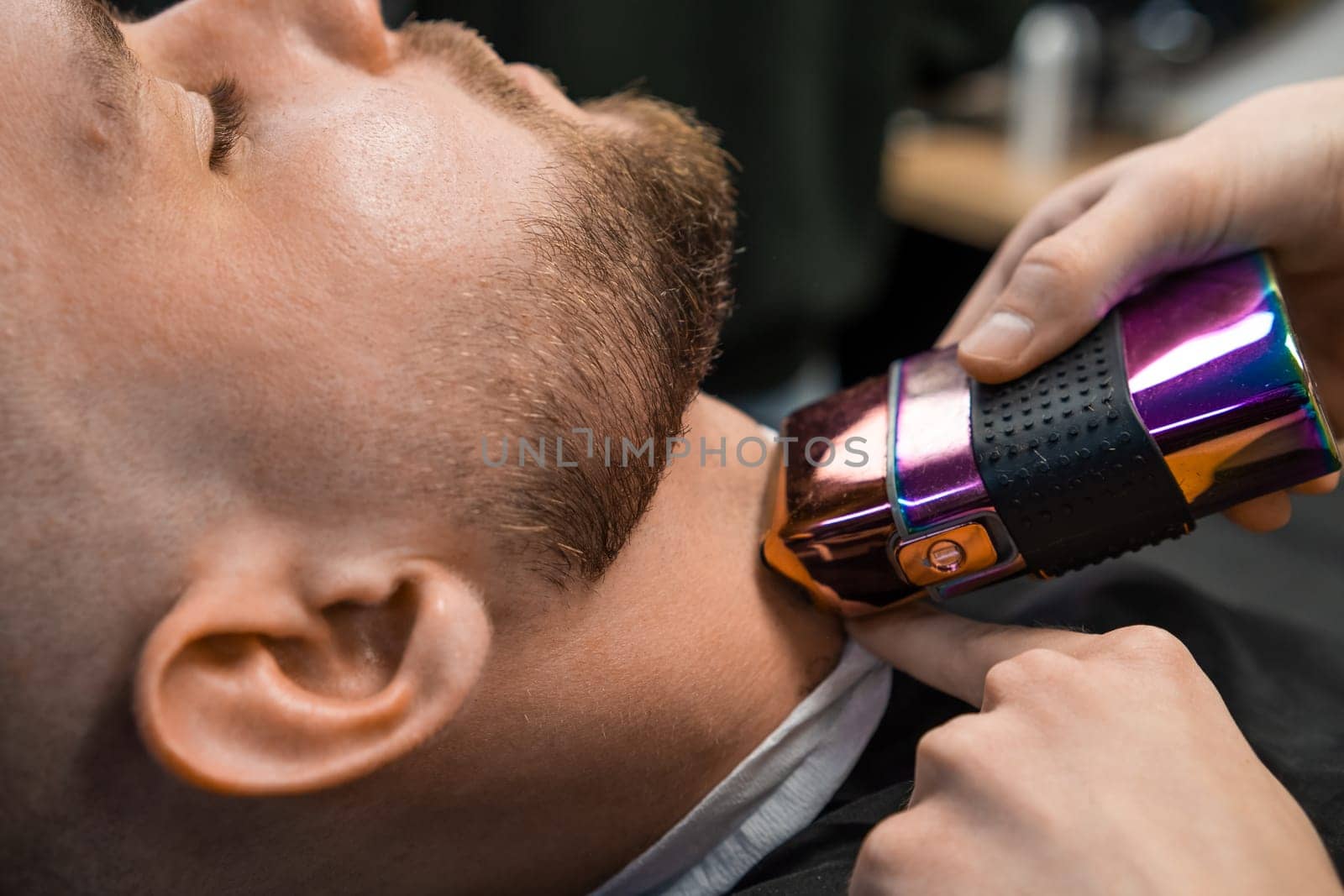 Master barber employs an automatic trimmer to groom the clients beard at the barbershop.