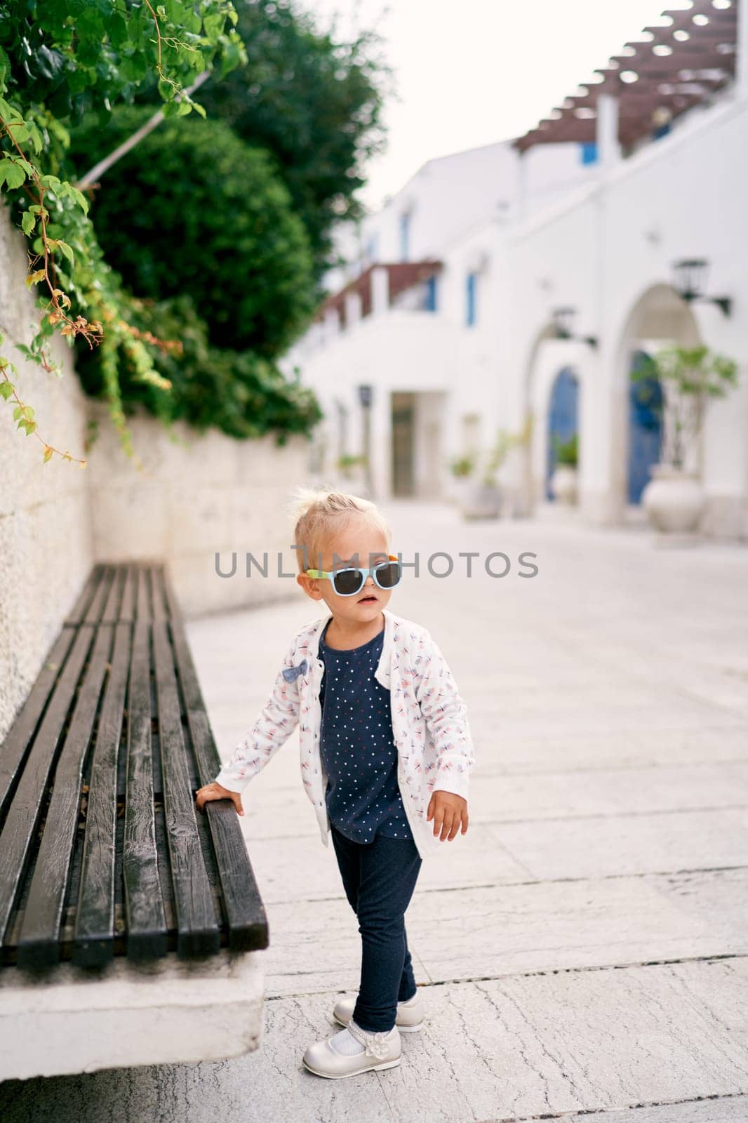 Little girl in sunglasses stands near a bench in the courtyard of the house, leaning on her hand. High quality photo