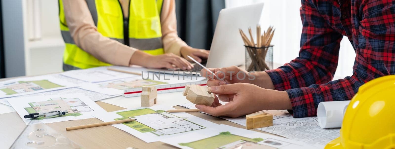 Professional architect and engineer collaborate to design eco-friendly house on meeting table with blueprint, wooden house block and tree model scatter around. Closeup. Delineation.