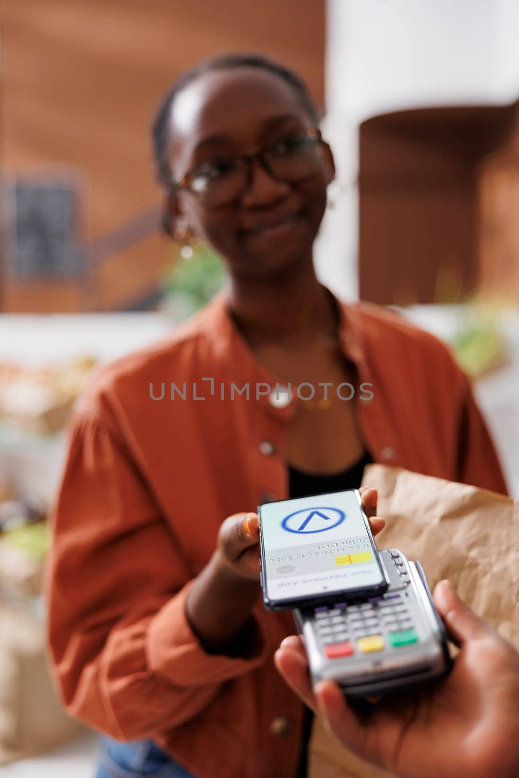 Detailed view of black individual grasping pos machine for female shopper to pay using her smartphone. Young customer utilizing her mobile device for nfc cashless payment at local grocery store.