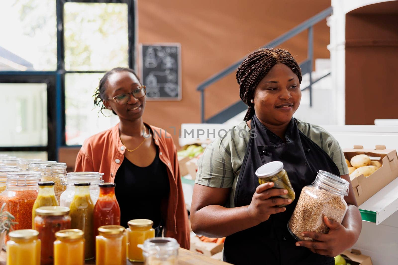 Female vendor and customer walking towards checkout counter with jars of preserved chemical free products. Black woman with an apron carrying bio organic items, assisting client at local store.