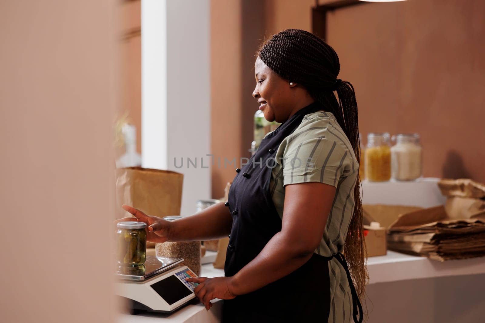 Side-view shot of african american female seller using weighing scale to measure glass container of fresh pickles. Black woman wearing an apron checking weight of organic product in a jar.