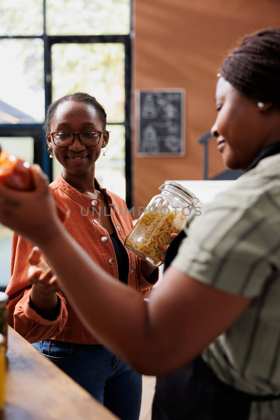 African American women hold organic products at bio supermarket, promoting sustainable, locally grown food. Female customer and vendor talking about health benefits of freshly made pasta and sauce.