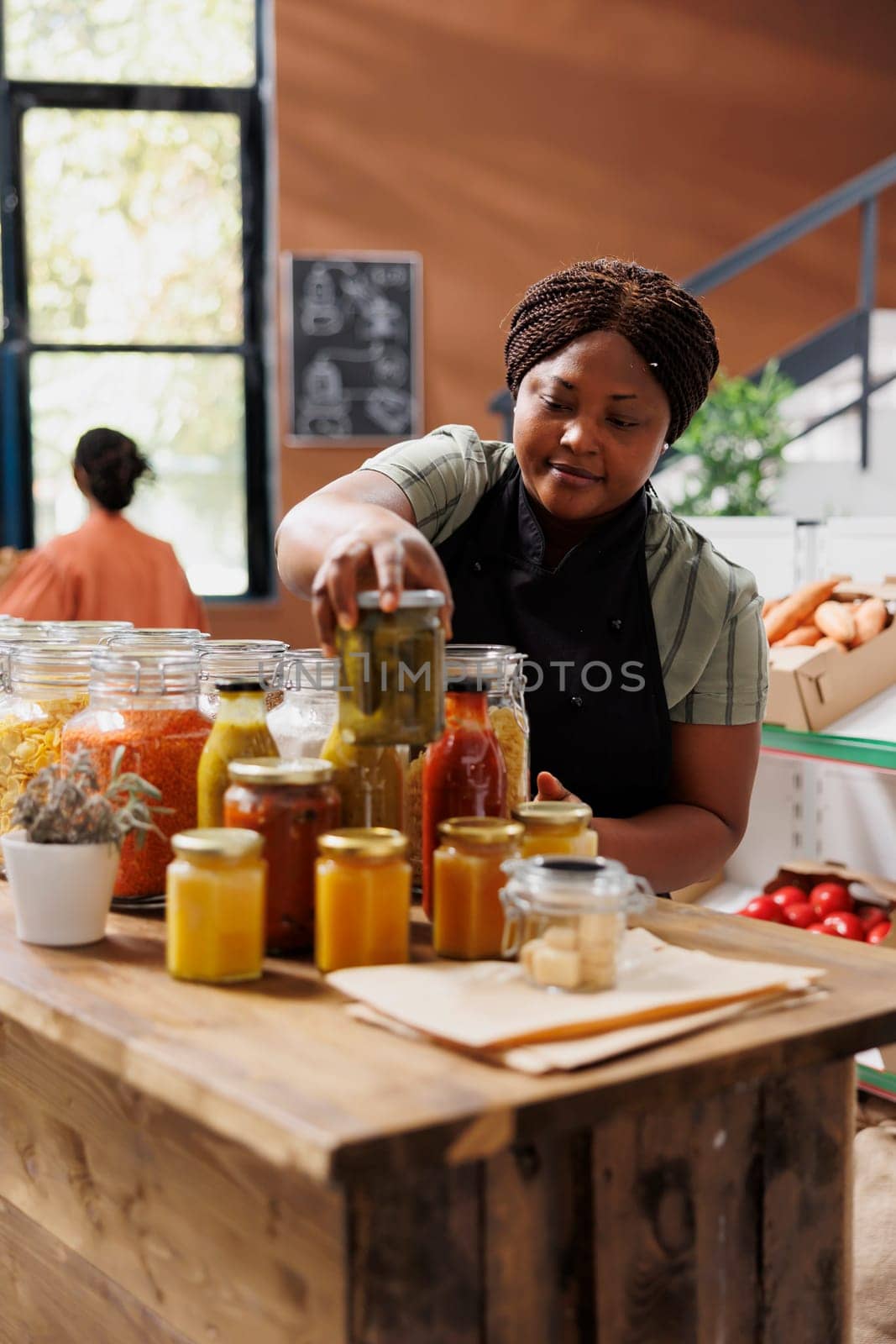 Female storekeeper selling jars of food, including pickles, in an environmentally conscious grocery shop. African american seller organizing reusable containers with organic preserved products.