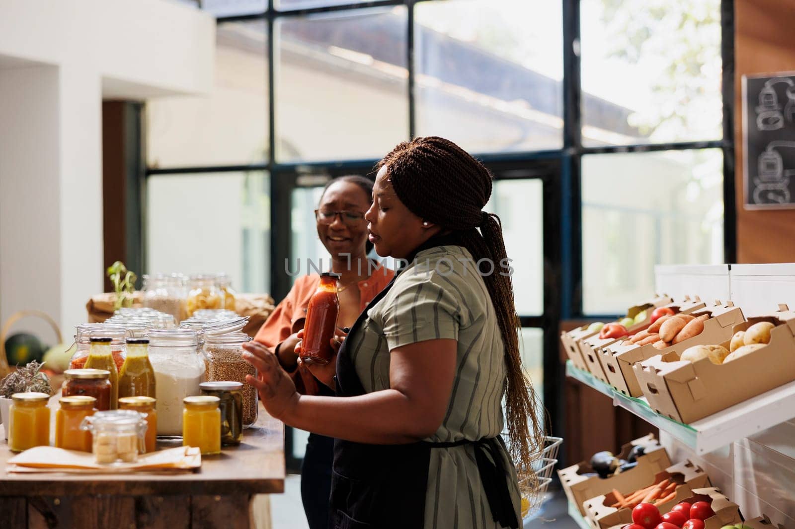Black woman with spectacles shopping in zero waste store looking for farm grown bulk products. Customer purchasing pantry staples from local grocery shop with assistance from female vendor.