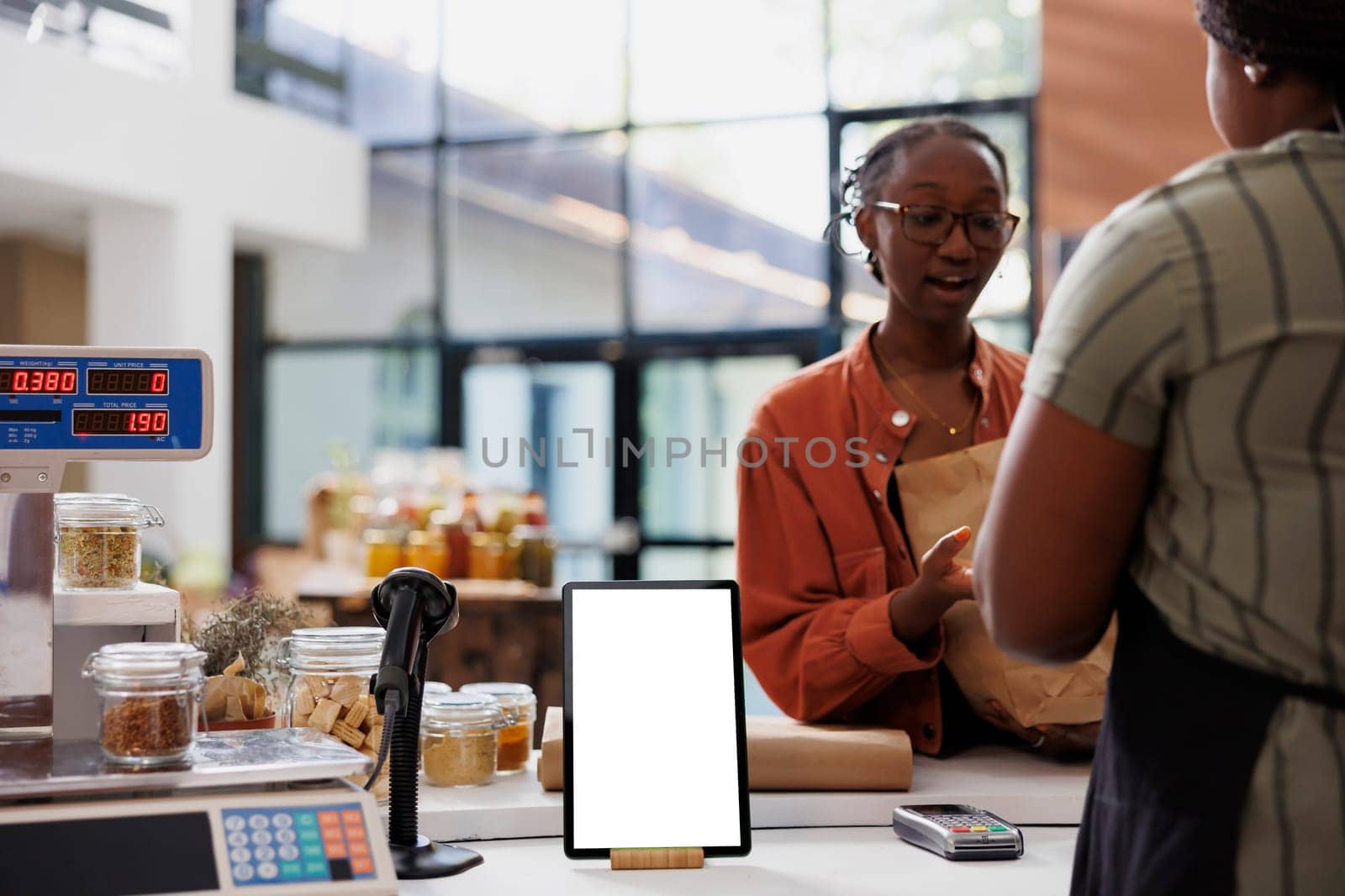 A lady wearing glasses converses with a vendor at the payment counter, while a digital device showcases a blank white screen featuring eco-friendly store advertisements. The tablet displays an empty chromakey mockup template