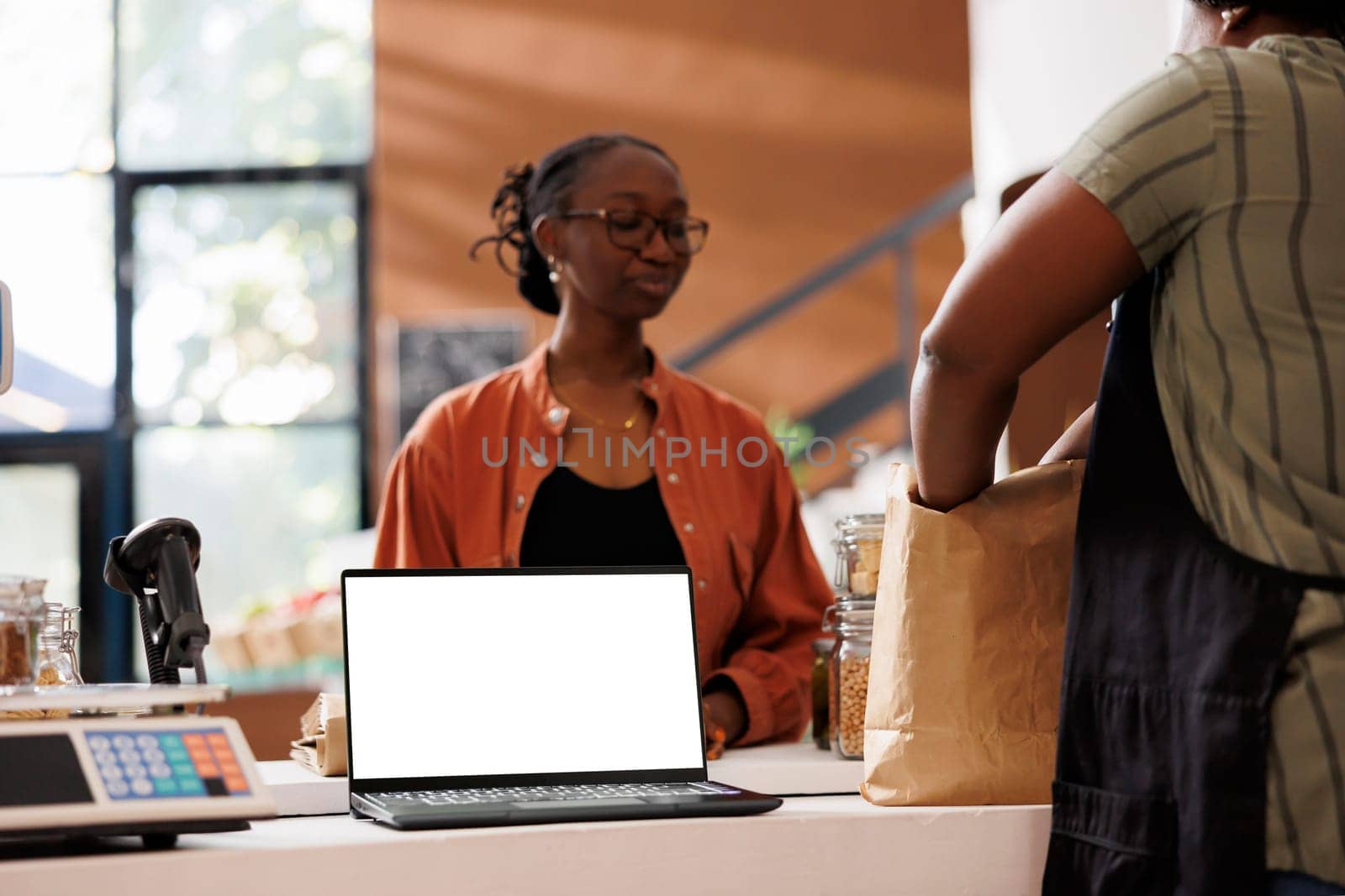 Seller places items in recyclable bags for the client who is waiting while laptop on counter displays an isolated white screen. Bio store uses mockup device with blank chromakey for commercials.