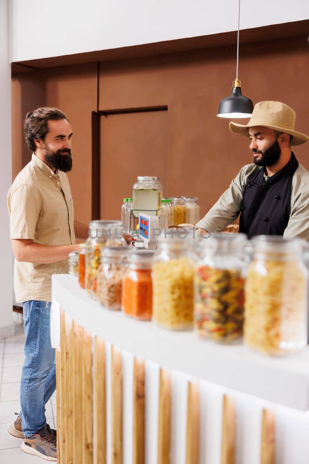 Young vendor stands at checkout counter of a zero waste shop, weighing farm grown cabbage for vegan customer with green lifestyle. Man shopping for groceries at an eco friendly store.