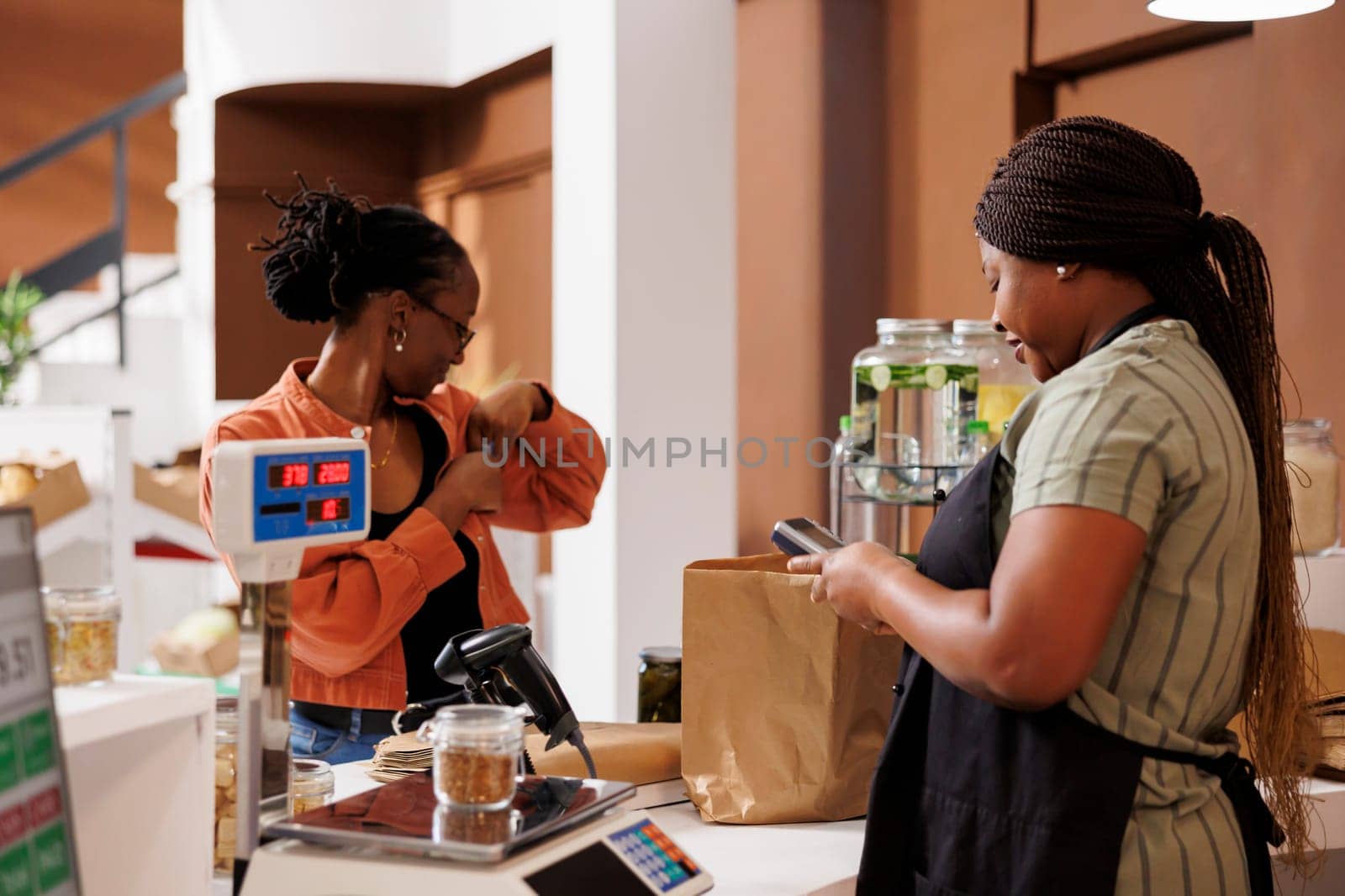 After paying for bio food items she selected at an eco friendly shop, black woman puts her credit card back in her pocket. At checkout counter, merchant and client complete a cashless transaction.