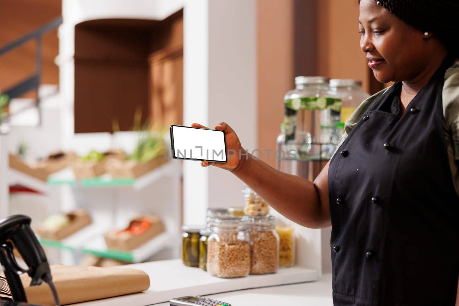 African American shopkeeper holds a mobile device horizontally that displays a white blank screen. Black woman with an apron is grasping smartphone showing isolated copy space mockup template.