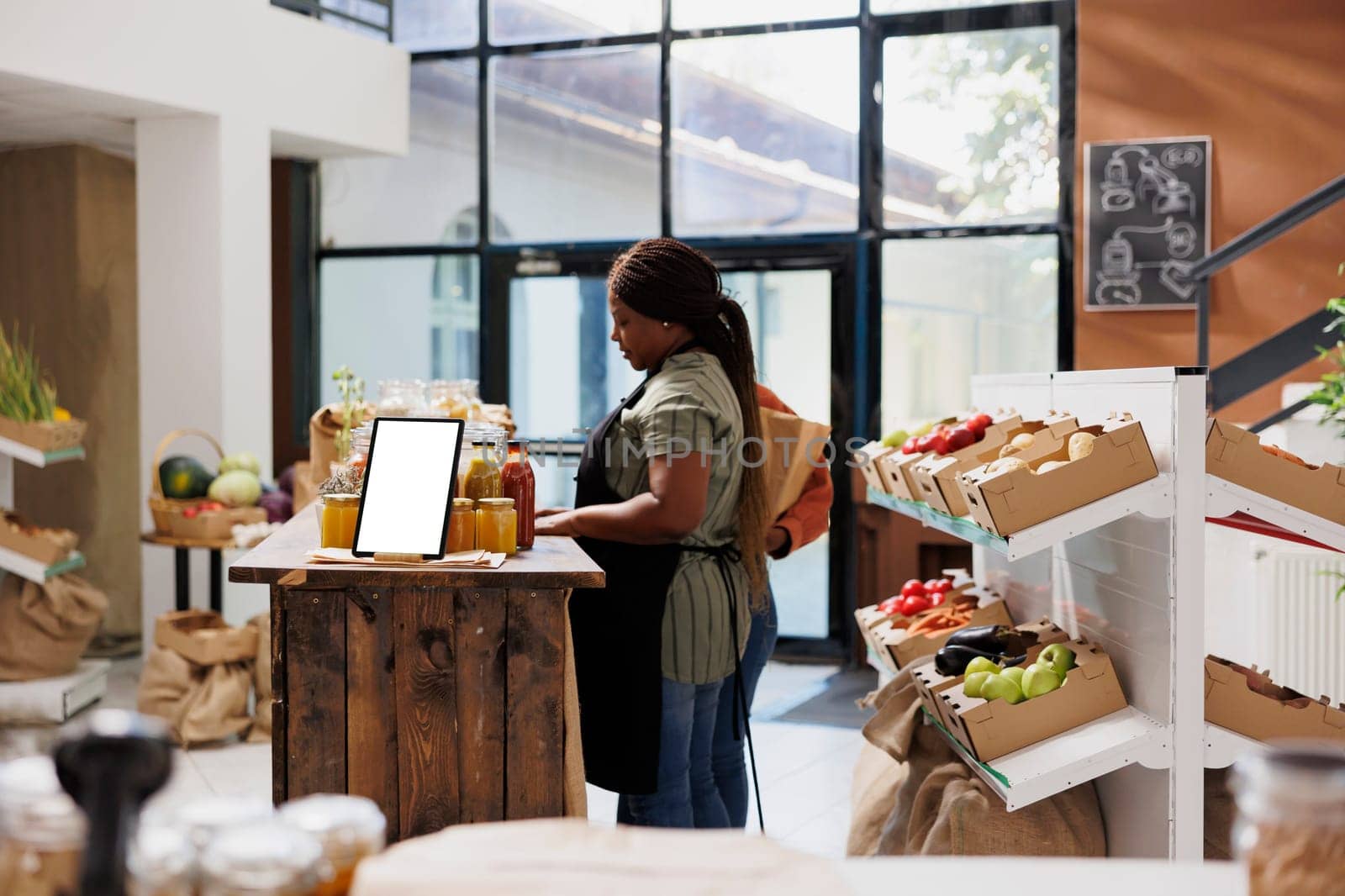 Vendor stands by table filled with glass jars filled with organic products while interacting with someone in environmentally friendly grocery store. Digital tablet on desk displaying white screen.