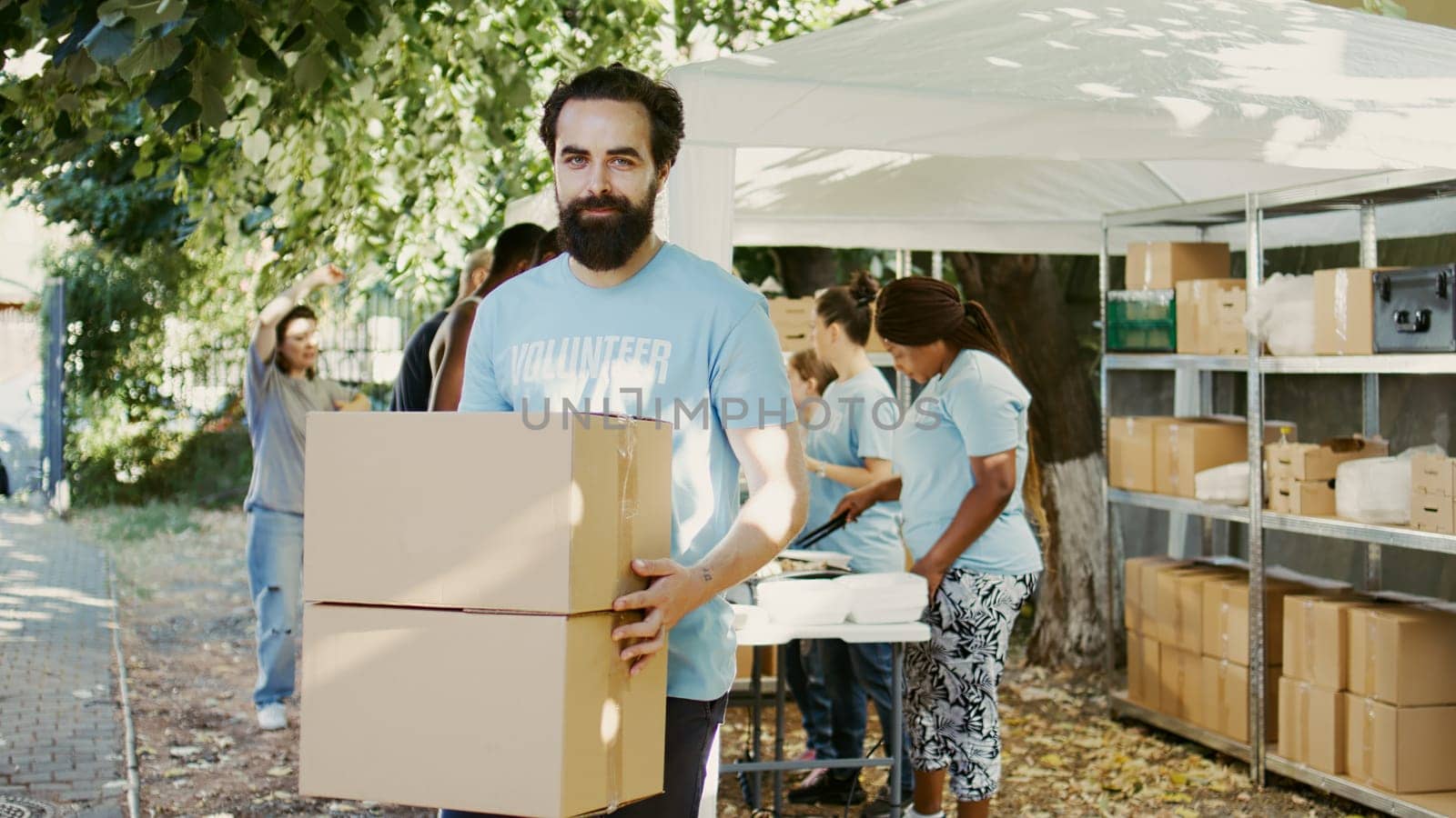 At a food drive, kind young guy holding non-perishable food products for the hungry and homeless. While glancing at the camera, male Caucasian volunteer carries donation packages. Handheld shot.