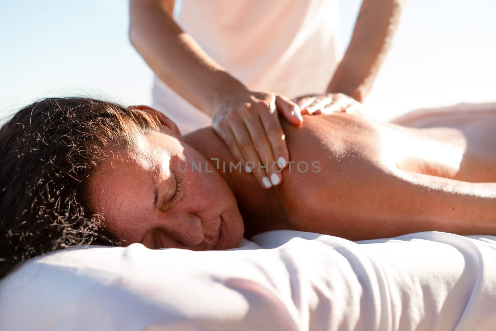 Crop anonymous masseur massaging the back of relaxed female tourist on special table. Brunette woman enjoying spa body treatment outdoors during summer vacation.