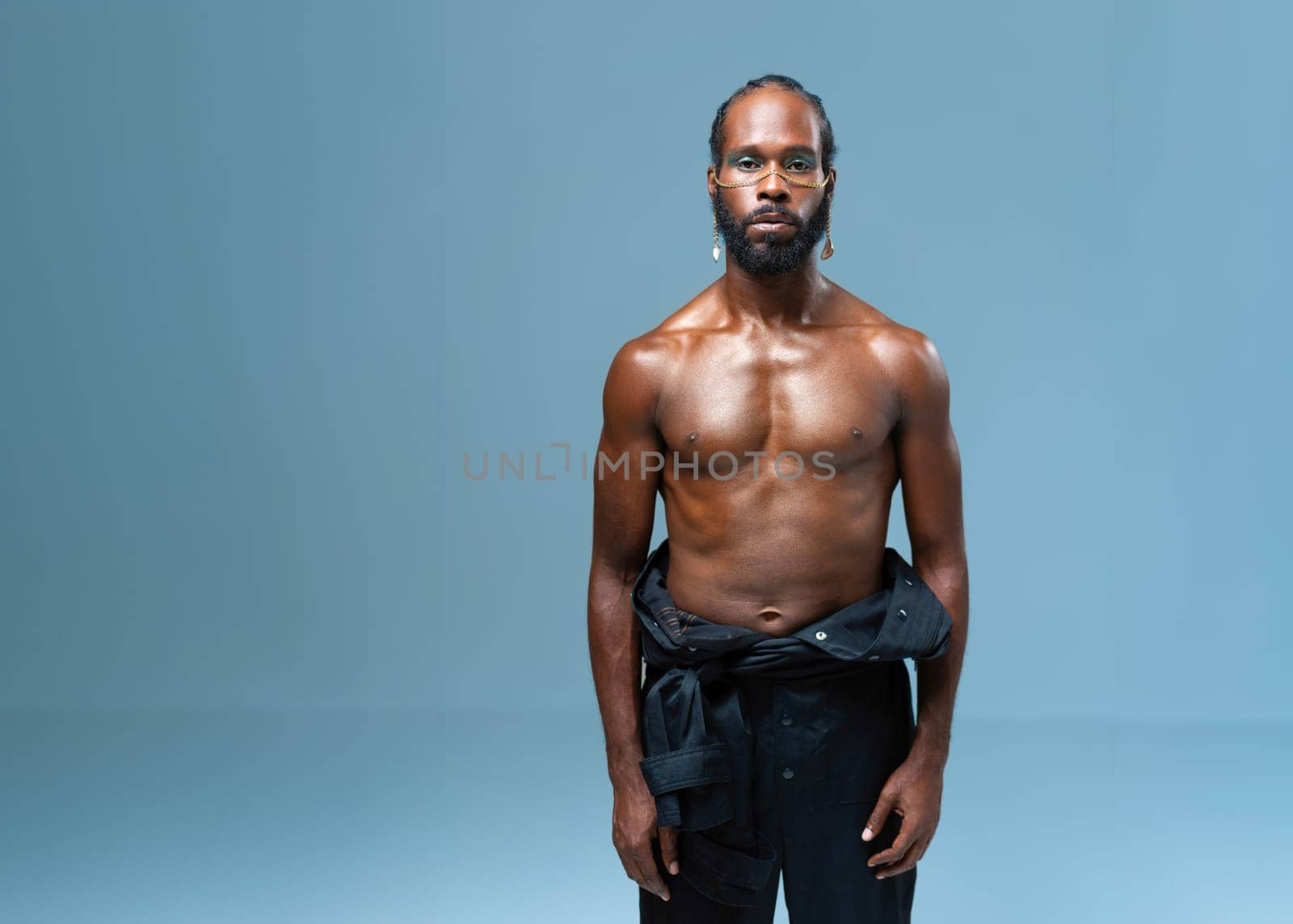 Portrait of shirtless African American gay man with light makeup standing isolated on blue background. Muscular black LGBTQ person in trendy pants looking at camera.