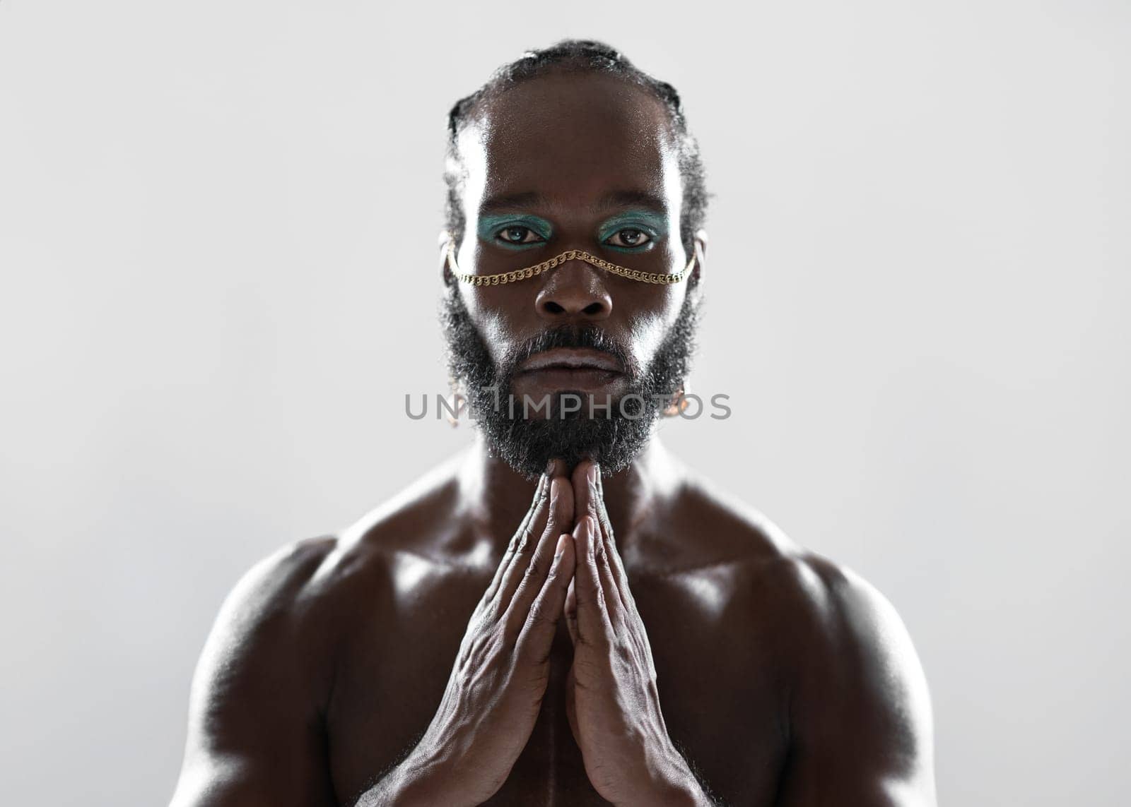 Confident African-American bearded gay man with bright makeup joining hands, isolated on white background. Close-up portrait of gay African man making namaste gesture and looking at camera.