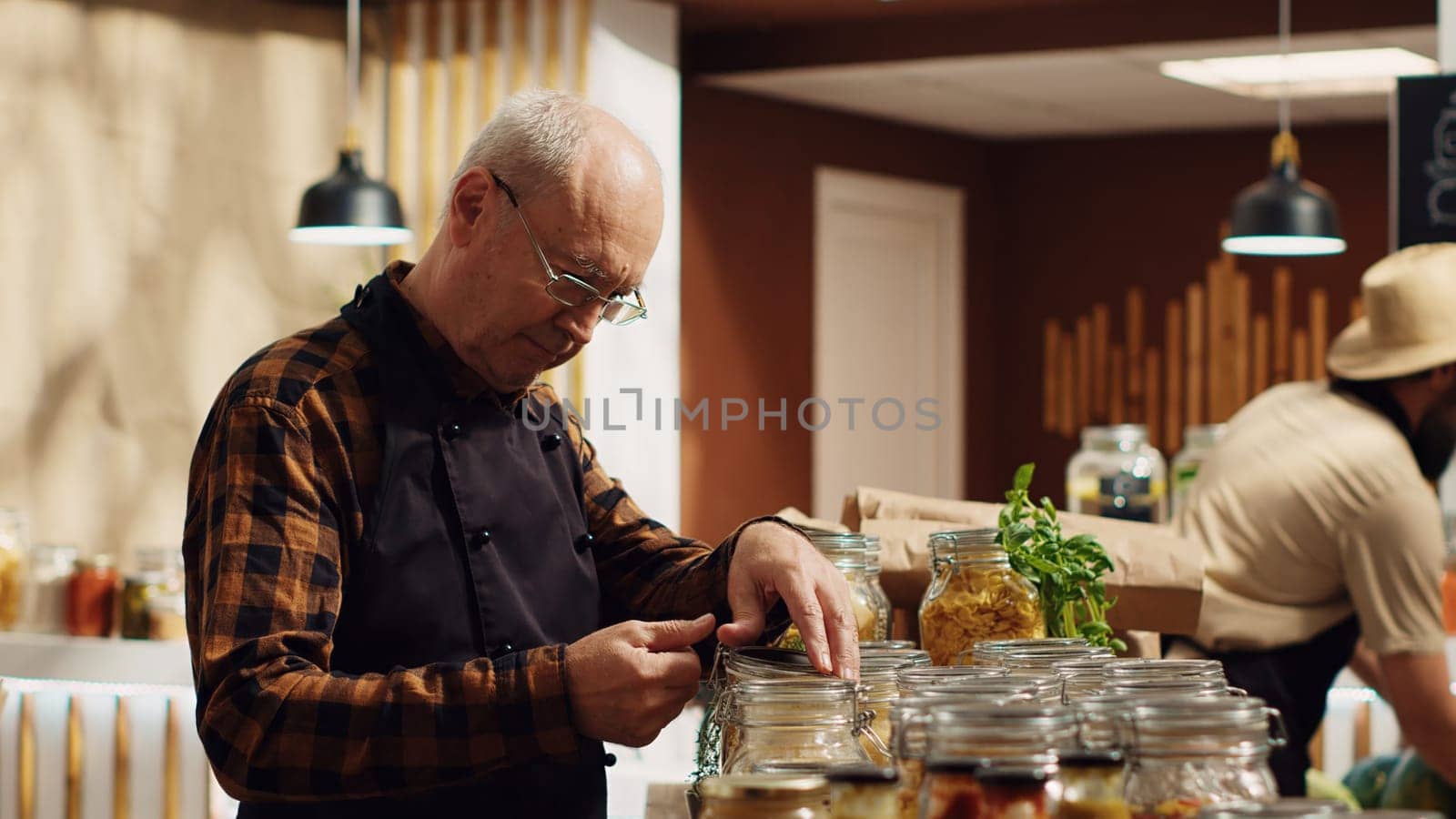 Portrait of smiling old zero waste vendor properly sealing bulk items decomposable glass containers. Elderly man in environmentally friendly local grocery shop arranging merchandise