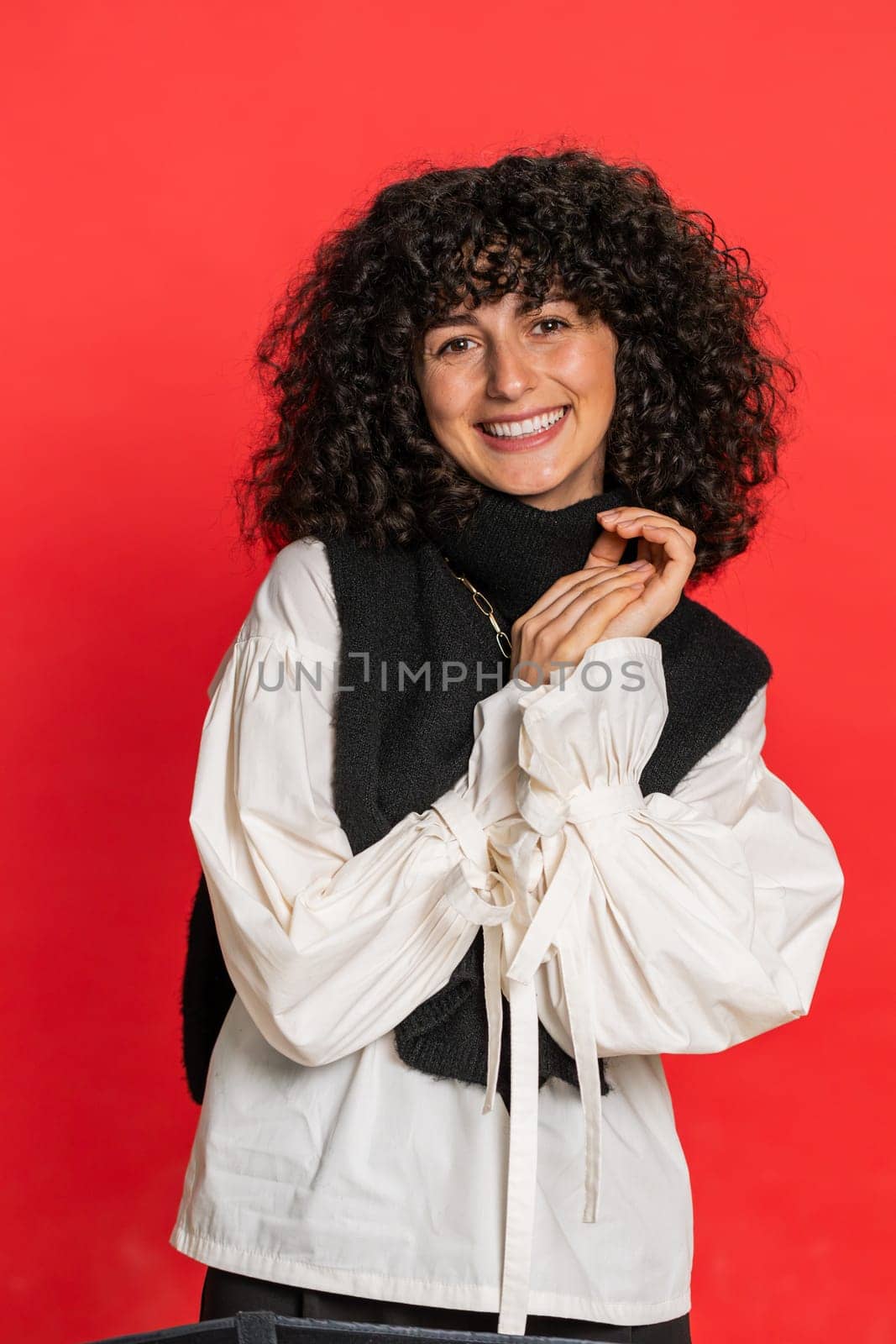 Portrait of happy Caucasian woman with curly hair smiling friendly, glad expression looking at camera dreaming, resting, relaxation feel satisfied good news. Girl indoors on red background. Vertical