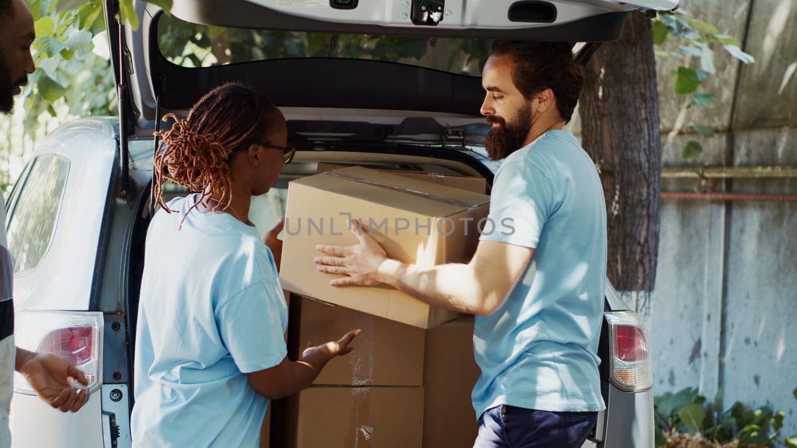 Male and female volunteers at the car giving charity boxes to poor needy and homeless people at community center. At food drive, the less privileged receive donations from a non-profit organization.