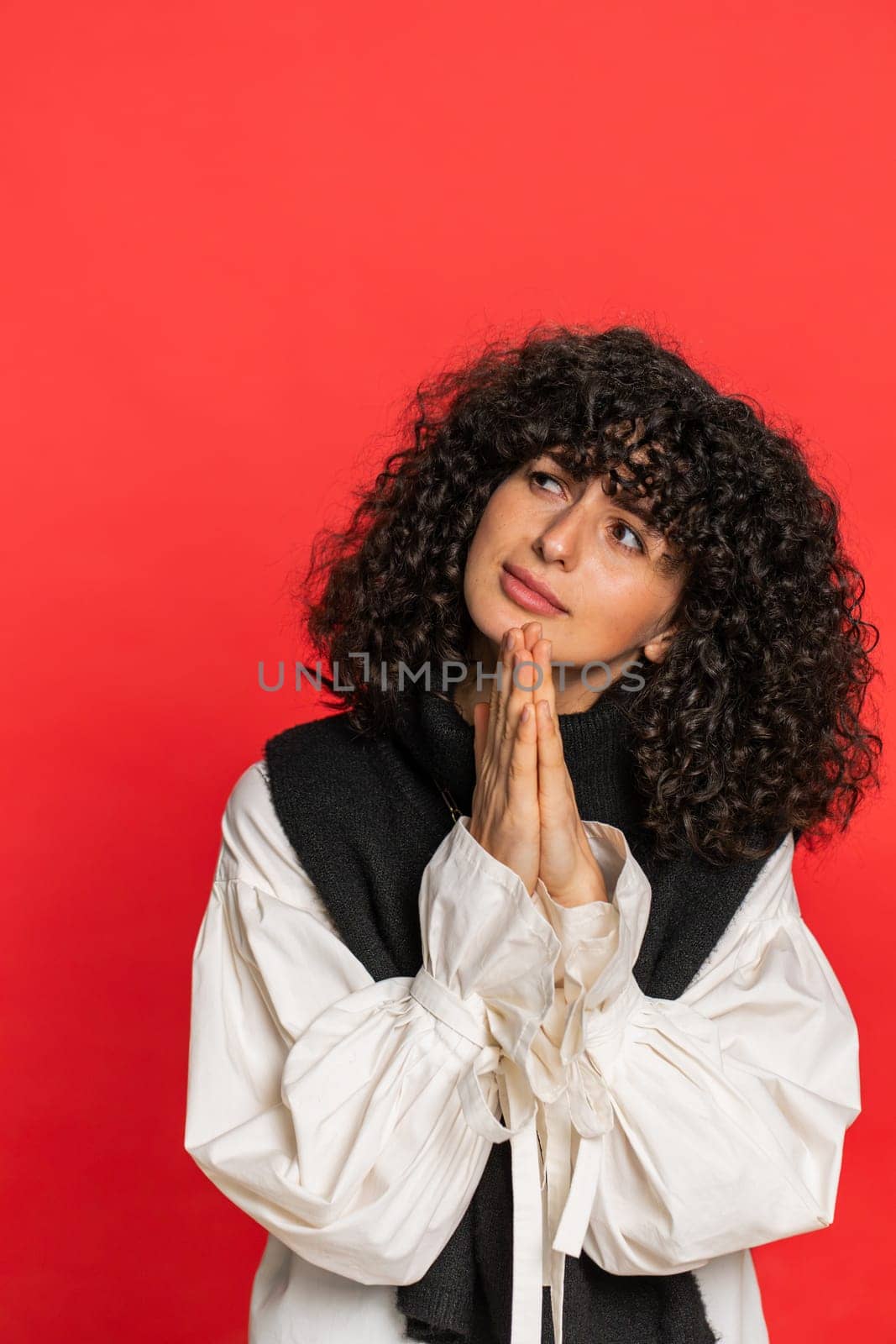 Please, God help. Young woman with curly hair praying, looking at camera and making wish, asking with hopeful imploring expression begging apology meditation. Girl isolated on red background. Vertical