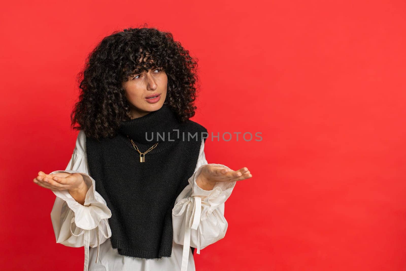 What. Why. Angry confused young woman with curly hair raising hands in indignant expression, asking reason of failure, demonstrating disbelief irritation by troubles. Girl isolated on red background