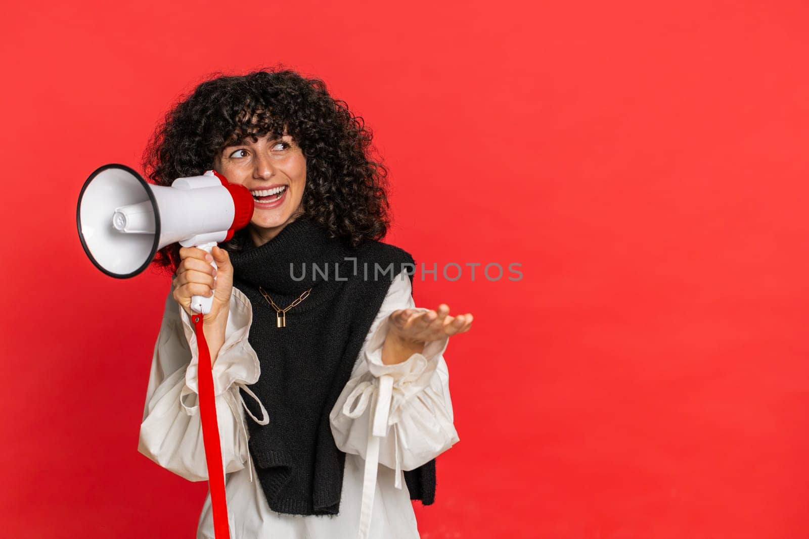 Caucasian young woman with curly hair talking with megaphone, proclaiming news, loudly announcing advertisement discounts sale, using loudspeaker to shout speech. Girl isolated on red background