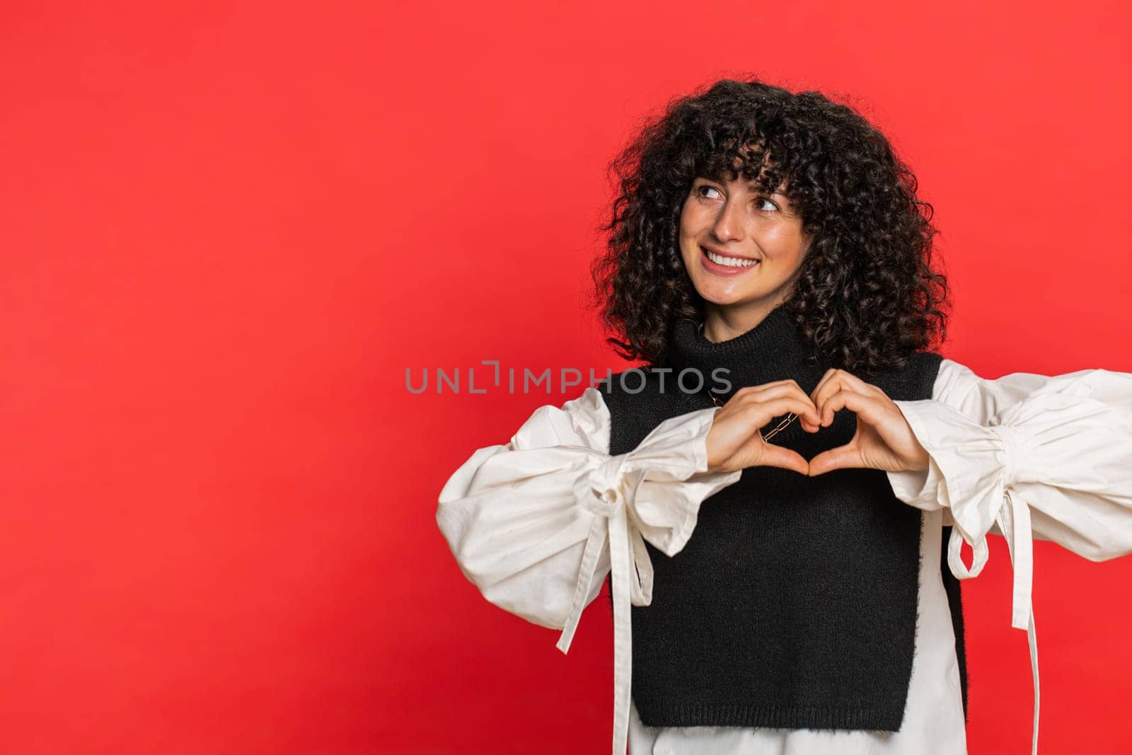 Woman in love. Smiling attractive Caucasian young woman with curly hair makes heart gesture demonstrates love sign expresses good positive feelings and sympathy. Girl isolated on red background