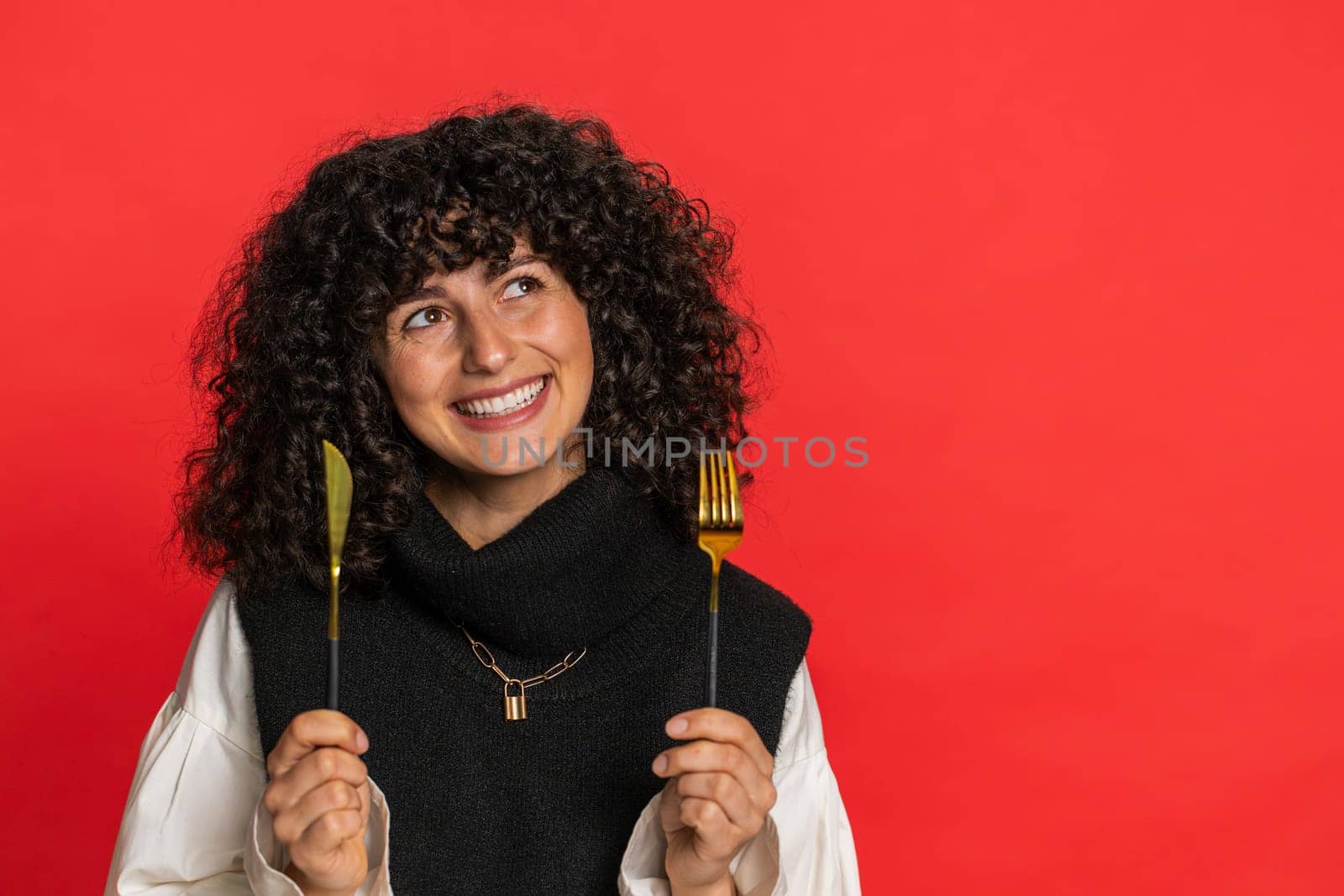 Ready to eat. Hungry Caucasian woman with curly hair waiting for serving dinner dishes with with restlessness holding cutlery fork knife, will appreciate delicious restaurant meal on red background