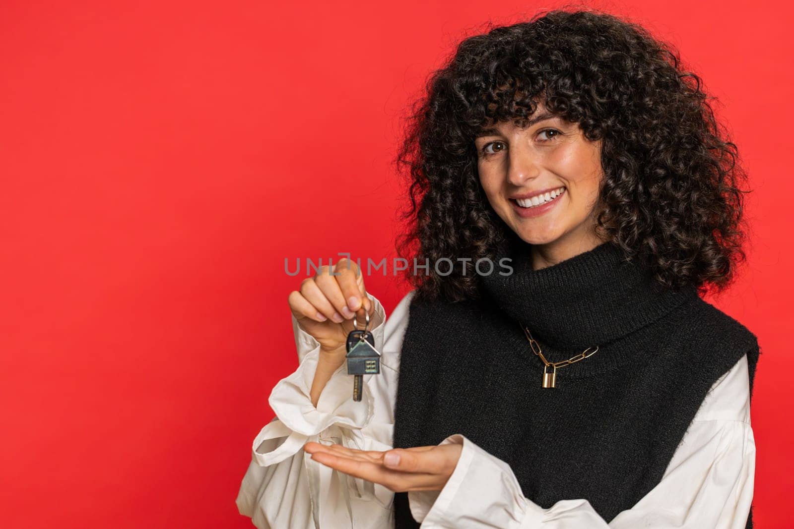 Caucasian young woman with curly hair real estate agent lifting hand showing the keys of new home house apartment, buying or renting property, mortgage loan, insurance. Girl isolated on red background