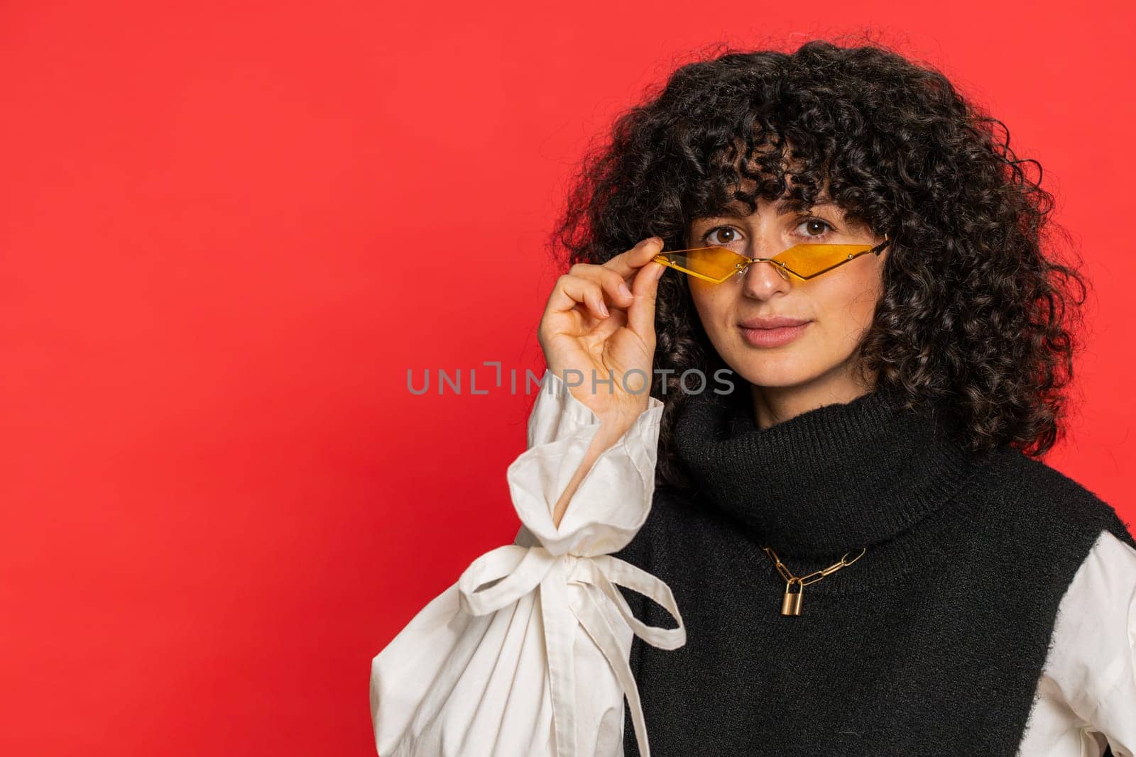 Stylish smiling Caucasian young woman with curly hair wearing orange sunglasses glad expression looking at camera dreaming, resting relaxation feel satisfied good news. Girl isolated on red background